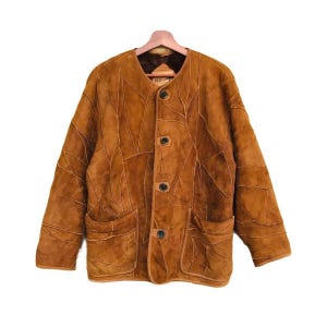 May include: A brown leather jacket with a patchwork design. The jacket has a round collar, four buttons, and two front pockets. The jacket is hanging on a wooden hanger.