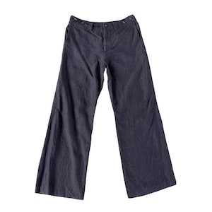 May include: Dark charcoal grey trousers featuring a wide-leg silhouette, a high-rise waistband with four visible buttons along the front closure, two side pockets, and a structured fly front design made from a textured fabric material.