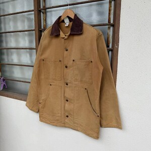 Puede incluir: Chaqueta de trabajo color canela con cuello de pana marr&oacute;n. La chaqueta tiene cuatro bolsillos delanteros y cierre de botones. Cuelga de una percha de madera. La chaqueta est&aacute; hecha de un material duradero y resistente.