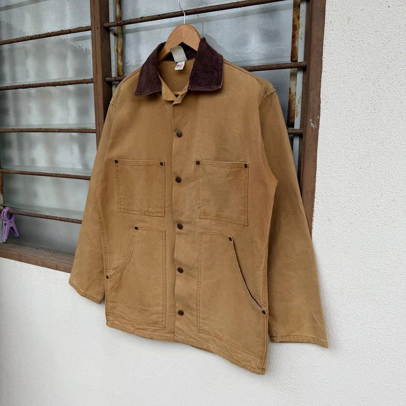 Puede incluir: Chaqueta de trabajo color canela con cuello de pana marr&oacute;n. La chaqueta tiene cuatro bolsillos delanteros y cierre de botones. Cuelga de una percha de madera. La chaqueta est&aacute; hecha de un material duradero y resistente.