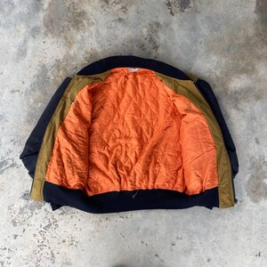 May include: A brown and black bomber jacket with an orange quilted lining. The jacket is unzipped and laid flat, showing the inside lining.