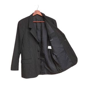 May include: Black pinstripe blazer with a classic design. The jacket features a notched lapel, three-button closure, and a pocket on each side. The interior lining is black, and the jacket is hanging on a wooden hanger.