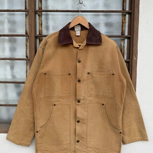 Puede incluir: Chaqueta de trabajo marr&oacute;n con cuello de pana marr&oacute;n, cuatro bolsillos delanteros y botones. La chaqueta est&aacute; colgada de una percha de madera contra una ventana.