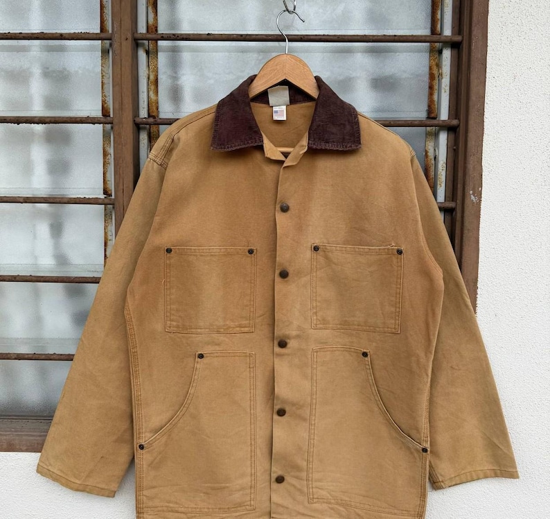 Puede incluir: Chaqueta de trabajo marr&oacute;n con cuello de pana marr&oacute;n, cuatro bolsillos delanteros y botones. La chaqueta est&aacute; colgada de una percha de madera contra una ventana.