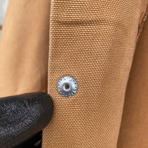 Puede incluir: Primer plano de una tela de color marr&oacute;n con un bot&oacute;n plateado. El bot&oacute;n es circular con un agujero central y est&aacute; sujeto a la tela. La tela tiene un tejido texturizado.