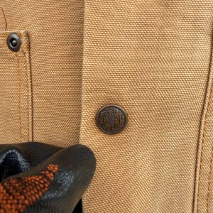 Puede incluir: Primer plano de una chaqueta de trabajo color beige con un bot&oacute;n met&aacute;lico con un dise&ntilde;o decorativo. La chaqueta tiene un bolsillo visible y detalles de costura. El bot&oacute;n es de color bronce oscuro.