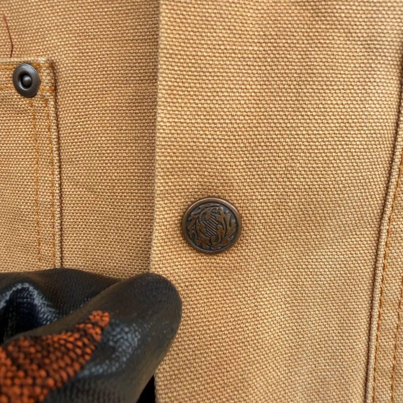 Puede incluir: Primer plano de una chaqueta de trabajo color beige con un bot&oacute;n met&aacute;lico con un dise&ntilde;o decorativo. La chaqueta tiene un bolsillo visible y detalles de costura. El bot&oacute;n es de color bronce oscuro.