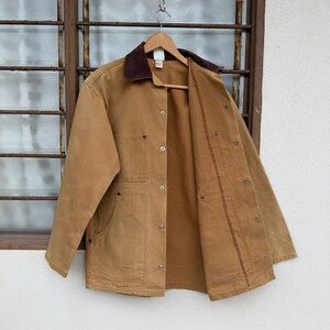 Puede incluir: Una chaqueta de trabajo color marr&oacute;n claro con cuello de pana marr&oacute;n cuelga de una percha de madera. La chaqueta tiene m&uacute;ltiples bolsillos, cierres de botones met&aacute;licos y un forro interior marr&oacute;n. La chaqueta se muestra contra una ventana.