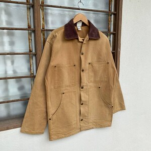 Puede incluir: Chaqueta de trabajo color canela con cuello de pana marr&oacute;n. La chaqueta tiene cuatro bolsillos delanteros, cierre de botones y est&aacute; colgada de una percha de madera. La chaqueta est&aacute; hecha de un material duradero.