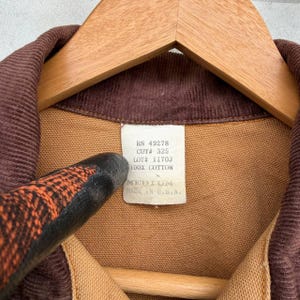 Puede incluir: Primer plano de un cuello de pana marr&oacute;n y una chaqueta de tela beige. La chaqueta est&aacute; colgada en una percha de madera. Una etiqueta blanca dice "RN 49278 CUT# 325 LOT# 1170J 100% COTTON MEDIUM MADE IN U.S.A."