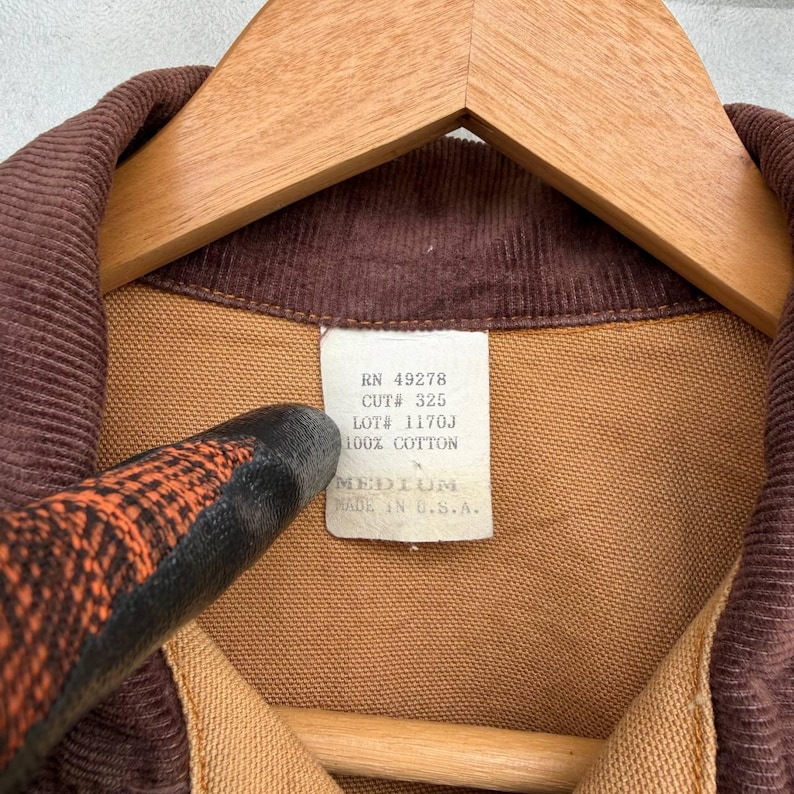 Puede incluir: Primer plano de un cuello de pana marr&oacute;n y una chaqueta de tela beige. La chaqueta est&aacute; colgada en una percha de madera. Una etiqueta blanca dice "RN 49278 CUT# 325 LOT# 1170J 100% COTTON MEDIUM MADE IN U.S.A."