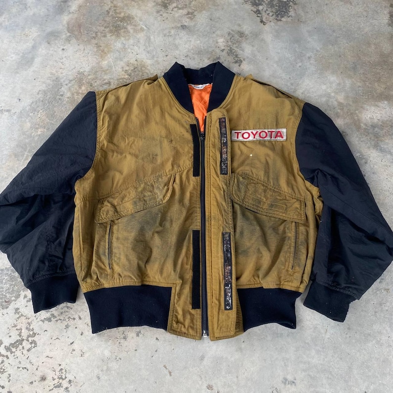 May include: A vintage brown and black bomber jacket with a zipper closure and a large "TOYOTA" patch on the right sleeve.