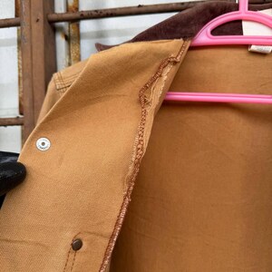 Puede incluir: Primer plano de una chaqueta de trabajo color canela con cuello de pana marr&oacute;n. La chaqueta tiene cierre de bot&oacute;n y detalle de costura. La chaqueta est&aacute; colgada en una percha rosa.
