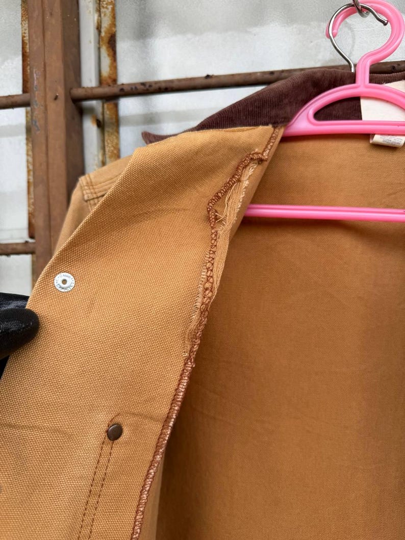Puede incluir: Primer plano de una chaqueta de trabajo color canela con cuello de pana marr&oacute;n. La chaqueta tiene cierre de bot&oacute;n y detalle de costura. La chaqueta est&aacute; colgada en una percha rosa.
