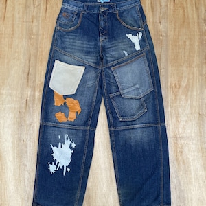 May include: Blue denim jeans with multiple patchwork details, including a white pocket, a brown leather patch, and a white paint splatter design.