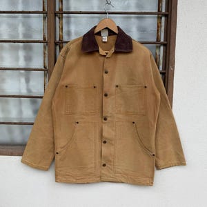 Puede incluir: Chaqueta de trabajo de color marr&oacute;n claro con cuello de pana marr&oacute;n, cuatro bolsillos delanteros y cierres de bot&oacute;n. La chaqueta est&aacute; colgada de una percha de madera frente a una ventana.