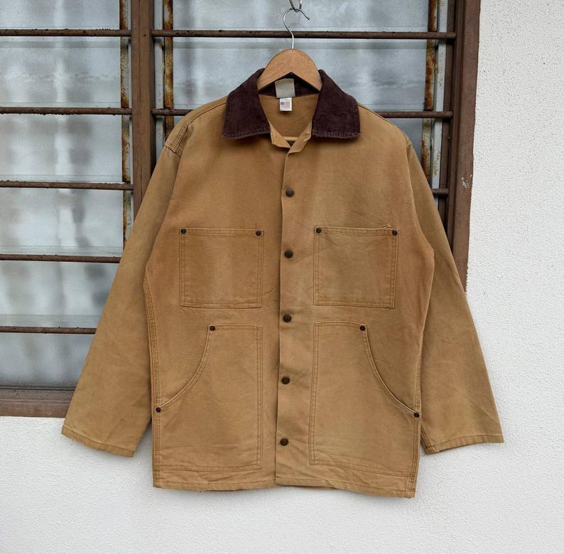 Puede incluir: Chaqueta de trabajo de color marr&oacute;n claro con cuello de pana marr&oacute;n, cuatro bolsillos delanteros y cierres de bot&oacute;n. La chaqueta est&aacute; colgada de una percha de madera frente a una ventana.