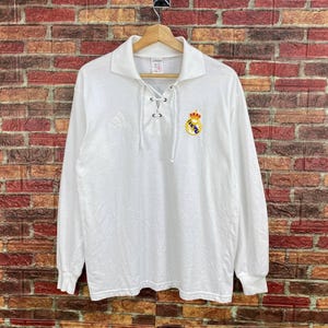 May include: White long-sleeve shirt with a collared neckline and lace-up detail. Features the Real Madrid crest on the chest. The Adidas logo is visible on the collar. The shirt is hanging on a wooden hanger against a brick wall.