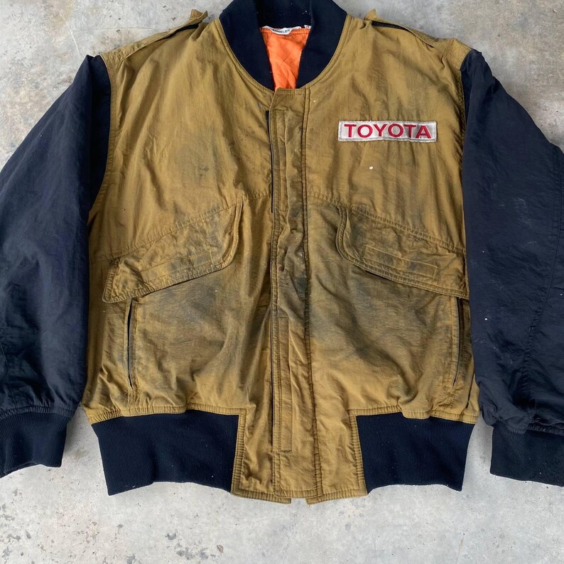 May include: A vintage brown and black bomber jacket with a large "TOYOTA" patch on the chest. The jacket has a zipper closure and two pockets on the front.