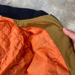 May include: Close-up of the inside of a brown and black bomber jacket. The lining is orange and quilted. The jacket has a brown collar and a black zipper.
