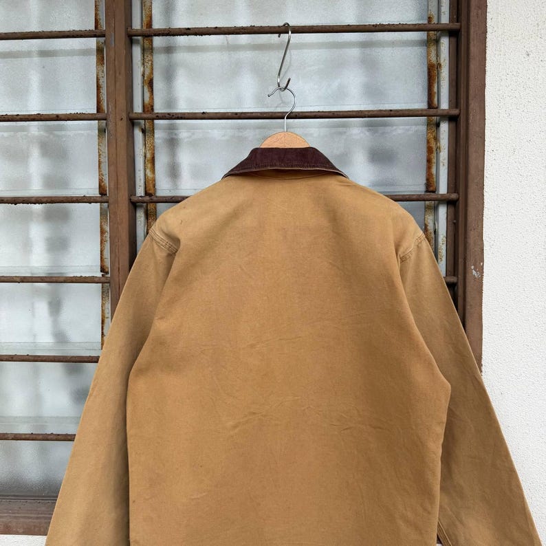 Puede incluir: Una chaqueta de trabajo color canela con cuello de pana marr&oacute;n cuelga de una percha de metal. La parte trasera de la chaqueta es visible, mostrando la tela duradera y el dise&ntilde;o cl&aacute;sico. La chaqueta se exhibe contra una ventana.