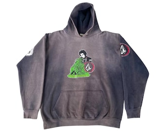 Può includere: Una felpa con cappuccio grigia con tasca frontale e cappuccio. La felpa presenta una grafica di una persona con fogliame verde e un logo rosso e bianco. Una toppa con il logo è sulla manica sinistra. La felpa è realizzata in materiale morbido.