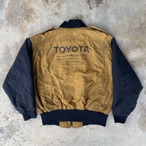 May include: A brown and black bomber jacket with a black zipper and black ribbed cuffs. The jacket has a large "TOYOTA" logo on the back with the text "Always Ahead of the Times" and "Highest Product Quality & Performance" underneath.