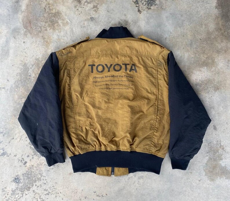 May include: A brown and black bomber jacket with a black zipper and black ribbed cuffs. The jacket has a large "TOYOTA" logo on the back with the text "Always Ahead of the Times" and "Highest Product Quality & Performance" underneath.