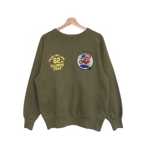 Puede incluir: Sudadera verde con un parche bordado en amarillo y azul de un oso de dibujos animados con un casco de piloto y sosteniendo una bandera. El texto "GRAVE INTL APT 62ND ILLINOIS USAF" está bordado en amarillo en la parte delantera de la sudadera.
