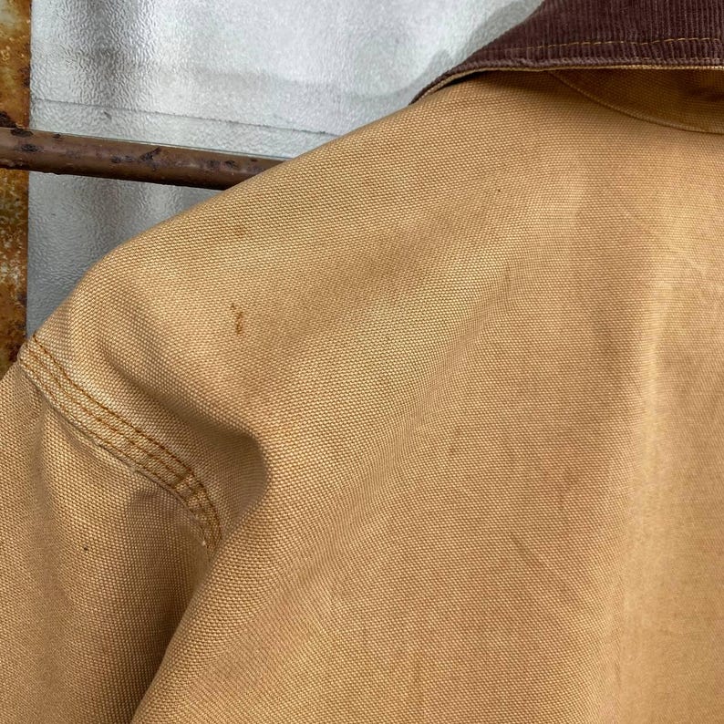 Puede incluir: Primer plano de una chaqueta de trabajo color beige con cuello de pana marr&oacute;n. La chaqueta presenta costuras visibles y algunas manchas oscuras. La tela parece ser de lona resistente, de estilo workwear.