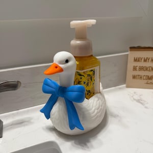 May include: A white ceramic goose-shaped soap dispenser with a blue bow around its neck. The goose has an orange beak and holds a yellow soap bottle. The dispenser is on a white countertop.
