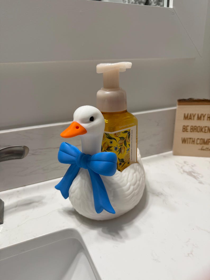 May include: A white ceramic goose-shaped soap dispenser with a blue bow around its neck. The goose has an orange beak and holds a yellow soap bottle. The dispenser is on a white countertop.