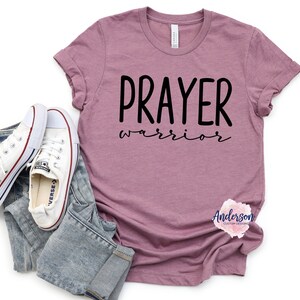 May include: A mauve t-shirt with the text "PRAYER warrior" in black lettering. The shirt is photographed on a white background with a pair of white sneakers and blue jeans.