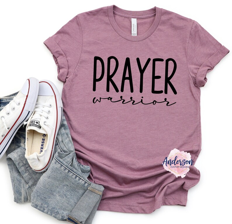 May include: A mauve t-shirt with the text "PRAYER warrior" in black lettering. The shirt is photographed on a white background with a pair of white sneakers and blue jeans.