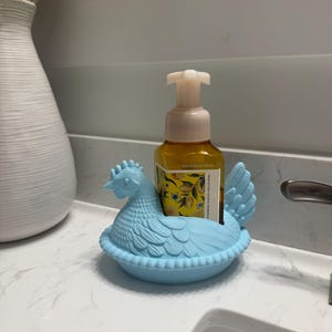 May include: A light blue chicken-shaped soap dispenser holder. The holder is made of a molded material and has a detailed design. A yellow soap bottle with a floral label sits inside the holder. The dispenser is on a white countertop.