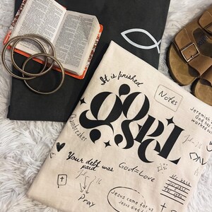 May include: A beige tote bag with black lettering that says "It is finished" and "God=Love". The bag also has a black and white illustration of a fish and the words "Chainbreaker", "Pray", "Jesus came for us", "Jesus died for us", and "He gives us peace".