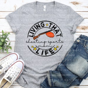 Puede incluir: Una camiseta gris con un dise&ntilde;o circular en blanco y negro que dice "Living That Shooting Sports Mom Life". El dise&ntilde;o incluye una escopeta de dibujos animados y una paloma de arcilla.