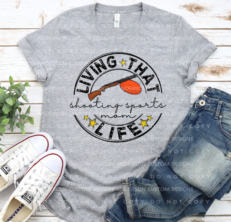 Puede incluir: Una camiseta gris con un dise&ntilde;o circular en blanco y negro que dice "Living That Shooting Sports Mom Life". El dise&ntilde;o incluye una escopeta de dibujos animados y una paloma de arcilla.