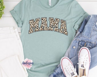 May include: A light blue t-shirt with a leopard print "MAMA" graphic. The shirt is laid flat on a white surface with a pair of white jeans, a denim jacket, and white sneakers.
