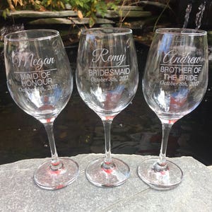 May include: Three clear glass wine glasses with personalized engravings. The glasses are engraved with the names "Megan", "Remy", and "Andrew" along with their respective titles and the date "October 8th, 2017".