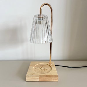 Puede incluir: Una lámpara de metal dorado con una pantalla de vidrio transparente y una base de madera. La base tiene un recorte circular con la letra "T" y el texto "Est. 2021" grabado en ella.