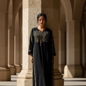 May include: A long black dress with floral embroidery on the neckline, sleeves, and sides. The dress has long sleeves and a tassel detail at the neckline. The background features stone columns and arches.