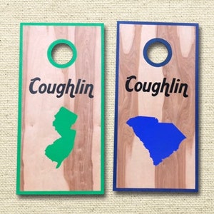 May include: Two wooden cornhole boards with the name "Coughlin" and a state outline. The board on the left has a green outline of New Jersey and the board on the right has a blue outline of South Carolina.