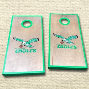 Könnte beinhalten: Zwei hölzerne Cornhole-Bretter mit grünem Rand und einem Philadelphia Eagles-Logo. Das Logo zeigt einen grünen Adler mit einem Football in seinen Krallen und die Worte "Philadelphia Eagles" in grün.
