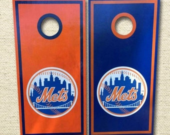 Puede incluir: Dos tableros de cornhole de madera con un logotipo de los New York Mets. Los tableros están pintados de naranja y azul con un círculo blanco alrededor del logotipo. El logotipo presenta el horizonte de la ciudad de Nueva York y la palabra "Mets".