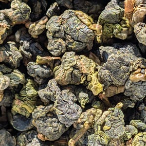 Könnte beinhalten: Nahaufnahme von getrockneten Teeblättern, die eine Mischung aus grünen, braunen und grauen Farbtönen zeigen. Die Blätter haben eine strukturierte, unregelmäßige Form, was auf ein hochwertiges, handverarbeitetes Produkt hindeutet. Das Bild hebt die natürlichen Farben und Details des Tees hervor.