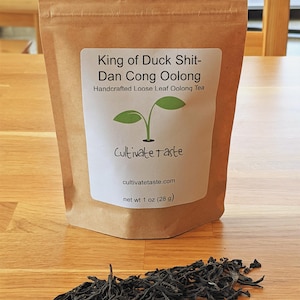 May include: A brown paper bag with a white label that reads "King of Duck Shit-Dan Cong Oolong Handcrafted Loose Leaf Oolong Tea Cultivate taste cultivatetaste.com net wt 1 oz (28 g)". A pile of loose leaf black tea is in front of the bag.