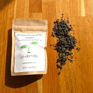 May include: A kraft paper bag of Ancient Snow Shan Green tea with a white label and green logo. The label reads "Handcrafted Loose Leaf Green Tea" and "cultivatetaste.com". Loose green tea leaves are scattered on a wooden surface. The bag contains 1 oz (28 g) of tea.