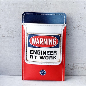 Peut inclure: Un étui rectangulaire de style pochette aux couleurs rouge, blanc et bleu avec des coutures blanches. L'avant présente un panneau rectangulaire avec un ovale rouge contenant le mot WARNING et le texte ENGINEER AT WORK en noir.
