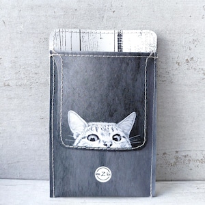 Peut inclure: Une pochette rectangulaire gris foncé avec une poche cousue présentant une illustration en noir et blanc d'un chat qui dépasse du haut. La pochette a un motif de grain de bois blanc sur le bord supérieur et un petit logo circulaire avec le texte bee Z loo.